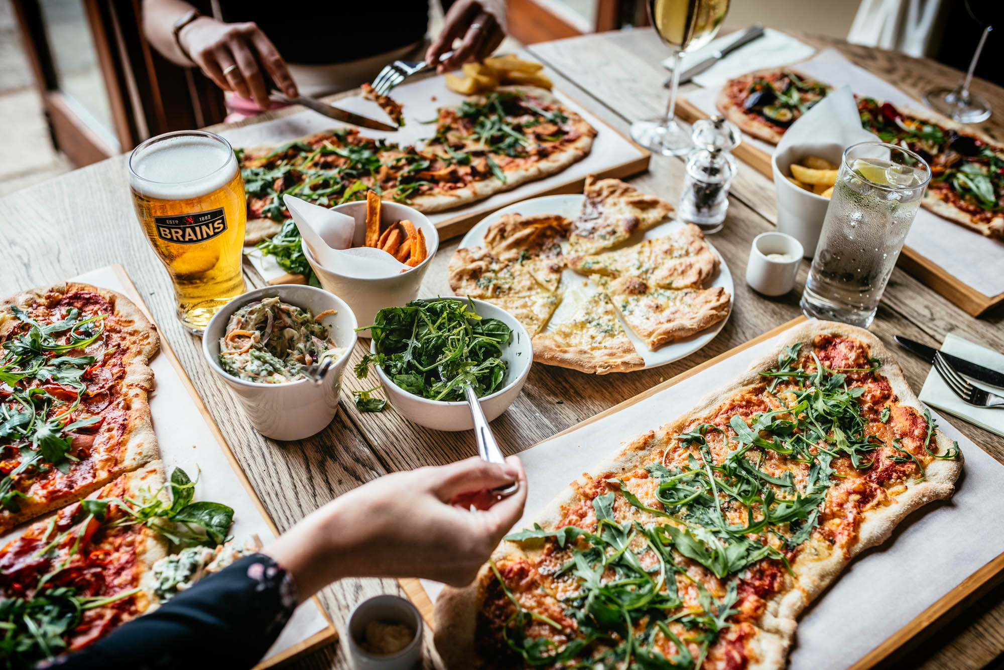 A photo of a table of food in a pub with four people sharing pizza, coleslaw, garlic bread and with drinks of beer, wine and sparkling water