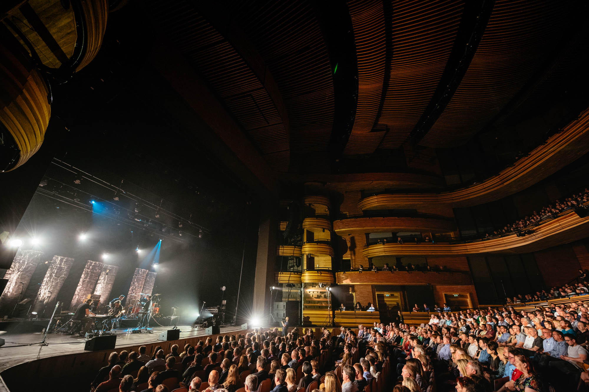 A photo of the inside of the Wales Millennium Centre Auditorium with a full house audience and stage Lighting