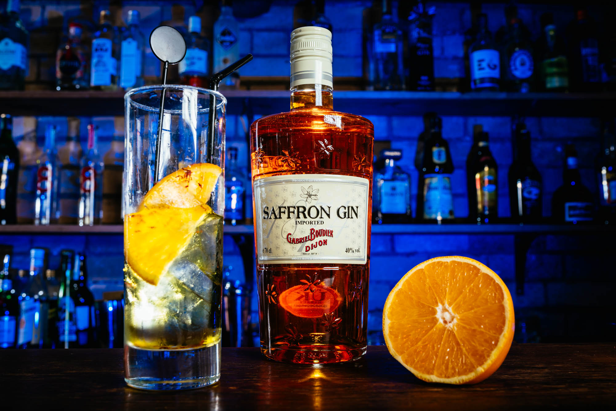 A photo of a bottle of saffron gin with a glass and a sliced orange in front of a blue lit bar