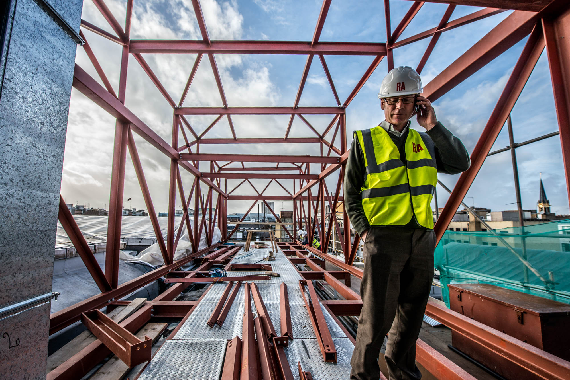 A photo of an manager engineer taking a phone call whilst stood in a building steel project with the london city skyline as the backdrop